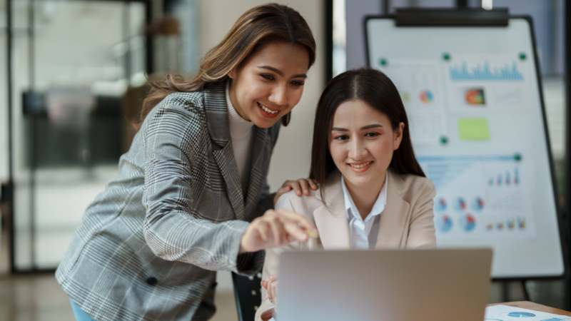 two female corporate executive one working on laptop and another standing behind her and pointing finger at laptop screen, both are smiling